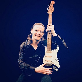 Photo of musician Simon Kinny-Lewis holding an electric guitar on their lap, guitar is pointing upwards with neck and end of guitar pointing towards the sky. Simon has long blond hair and is wearing all black